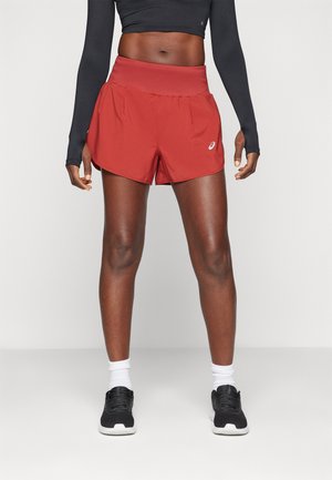 Red running shorts with a wide waistband, pleated design, and a logo detail on the left. Worn with a black long-sleeve top and black sneakers.