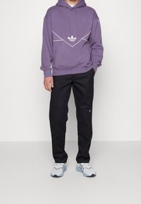 Lila hoodie med vitt geometriskt mönster och känguruficka, kombinerad med svarta byxor med ett märke, bärs med ljusblå sneakers.