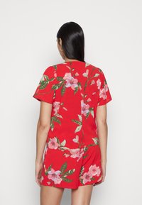 Macacão floral vermelho de mangas curtas, com padrões de flores rosa e verde. Tecido suave com um corte relaxado e um simples decote redondo.