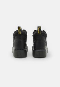 As botas de tornozelo pretas apresentam um cabedal texturizado com um painel em malha e uma sola espessa de borracha. As abas traseiras têm o texto amarelo "Bouncing Soles".