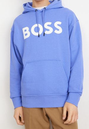 Person trägt einen blauen Hoodie mit weißem "BOSS" Text über der Brust und braune Hose, Hände entspannt an den Seiten.
