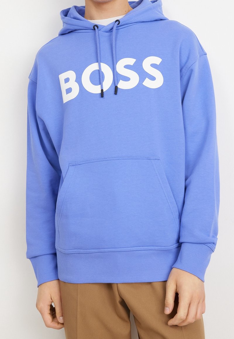 Personne portant un sweat à capuche bleu avec un texte blanc "BOSS" sur la poitrine et un pantalon marron, mains détendues le long du corps.