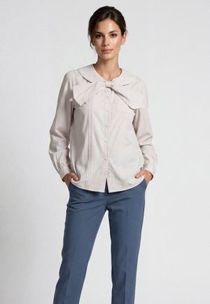 Frau trägt eine hellbeige Bluse mit großer Schleife am Kragen und blaue, maßgeschneiderte Hose, steht mit den Händen in den Taschen.