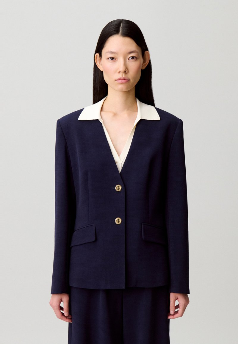 Blazer blu navy con design strutturato, collo bianco, bottoni dorati e due tasche frontali, realizzato in un tessuto liscio e leggero.