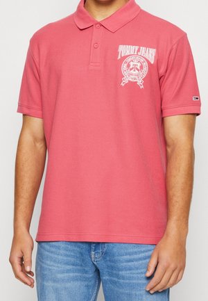 Pink polo-shirt lavet af tekstureret stof, med et hvidt logo-design på venstre bryst og standard krave. Bæres med lyseblå jeans.