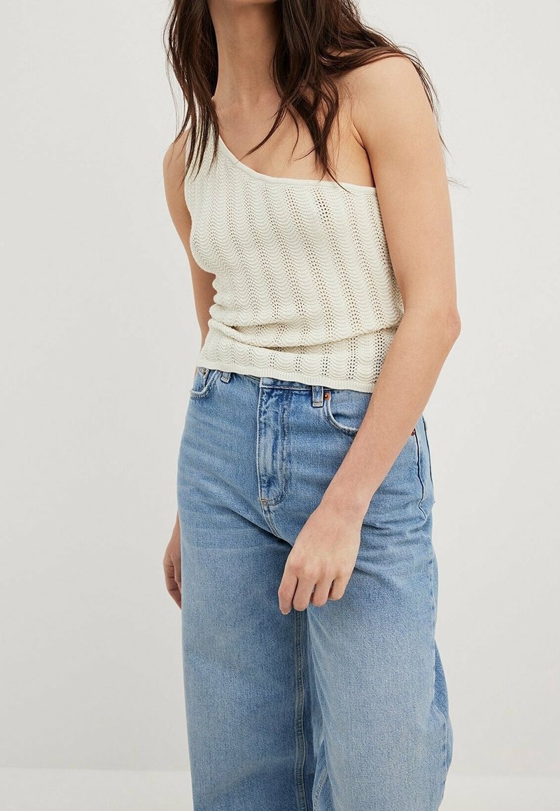 Femme portant un top en maille crème à une épaule et un jean large bleu clair taille haute, debout devant un fond uni.