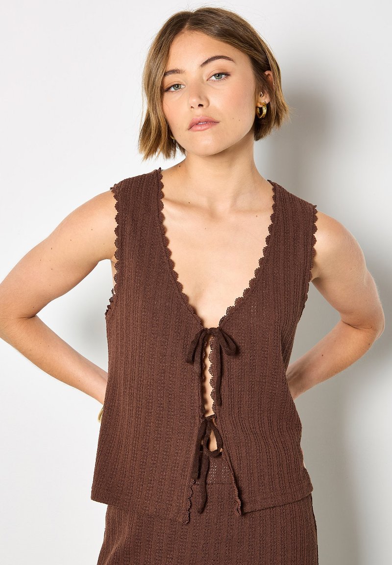 Femme portant un haut marron sans manches en maille avec des bords festonnés et des liens à nouer, associé à un bas en maille assorti, debout contre un mur blanc.