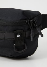 Černá nylonová ledvinka má zipovou hlavní kapsu, nastavitelný popruh a texturované akcenty. Na přední straně je výrazná nášivka s logem.