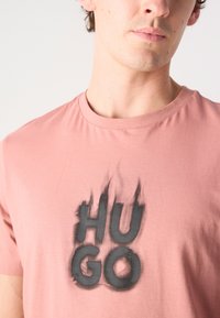 Rozā kokvilnas T-krekls ar melnu liesmu stila grafiku un tekstu "HUGO." Apaļa kakla izgriezums, īsas piedurknes un gluda tekstūra.