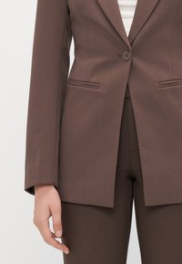 Hnědý upravený blazer se zapínáním na jeden knoflík, dvěma předními kapsami, strukturovanými rameny a rovním lemem. Látka se zdá být hladká a texturovaná.