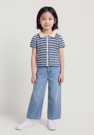 Jeune fille debout, portant un haut à manches courtes boutonné à rayures bleu et crème, un jean large bleu clair, et des chaussures blanches sans lacets.