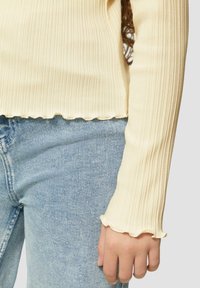 Crème ribbeltrui met lange mouwen en golvende zoom; gedragen met lichtblauwe denim jeans. Close-up toont de gestructureerde stof en de aansluitende pasvorm.