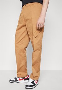 Tan cargobyxor med fickor, en avslappnad passform och knappstängning. Bärs med svart- och vitfärgade sneakers som har röda detaljer.