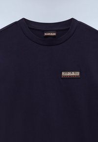 Marineblå bomulds t-shirt med rund hals og et lille rektangulært logo patch med hvid og rød tekst på brystet. Glat tekstur.