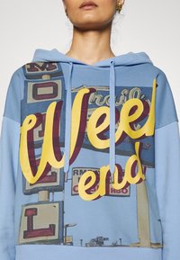 Niebieska bluza z kapturem z dużym nadrukiem graficznym z tekstem "Wee kend" w kolorze żółtym i burgundowym, z tłem przedstawiającym oznakowanie motelu.