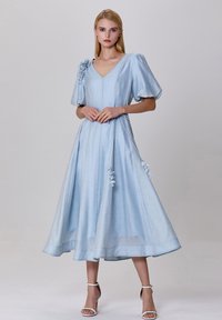 Robe bleu clair, longueur genou en un tissu doux et texturé. Elle présente des manches bouffantes, un col en V et des accents floraux décoratifs sur les côtés.