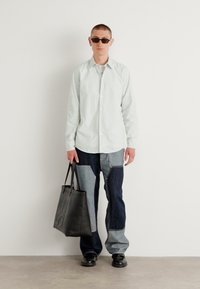 Camisa de botón verde claro, pantalones de mezclilla azul en dos tonos con diseño de parches, bolsa de mano negra, zapatos negros, gafas de sol, entorno minimalista.