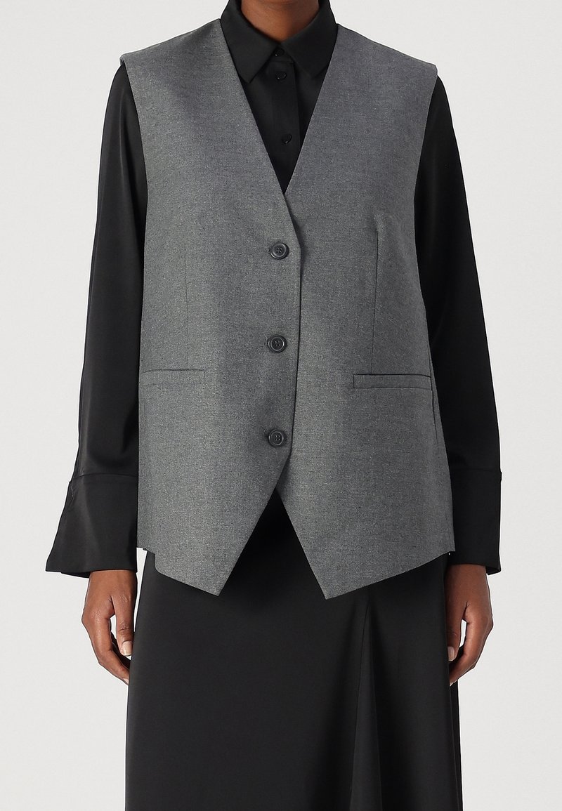 Personne portant un gilet gris ajusté boutonné par-dessus une chemise noire à manches longues avec de larges poignets et une jupe noire.