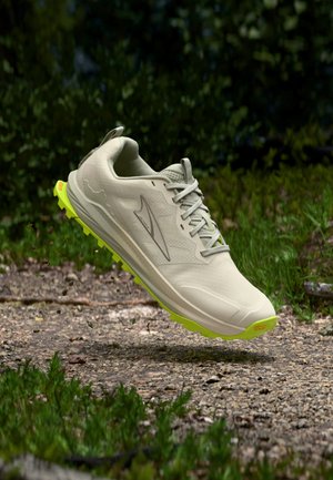 Bēša taku skriešanas kurpe ar dzeltenu Vibram zoli, sasietām šņorēm, gaisā virs grants takas ar zaļumu fonā.