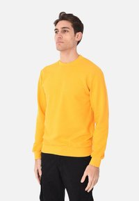 Felpa gialla brillante con collo rotondo e maniche lunghe, realizzata in tessuto morbido, con polsini e orlo a costine. Indossata con pantaloni neri.