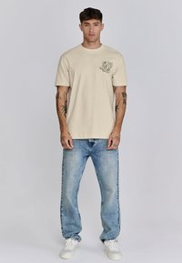 Beige Baumwoll-T-Shirt mit kurzen Ärmeln, das ein schwarzes Logo auf der linken Brust trägt. Kombiniert mit hellblauen Jeans und weißen Sneakers.
