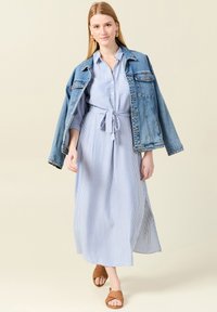 Abito lungo a righe blu e bianche con vita annodata, abbinato a un giubbotto in denim strappato e sandali intrecciati marroni.