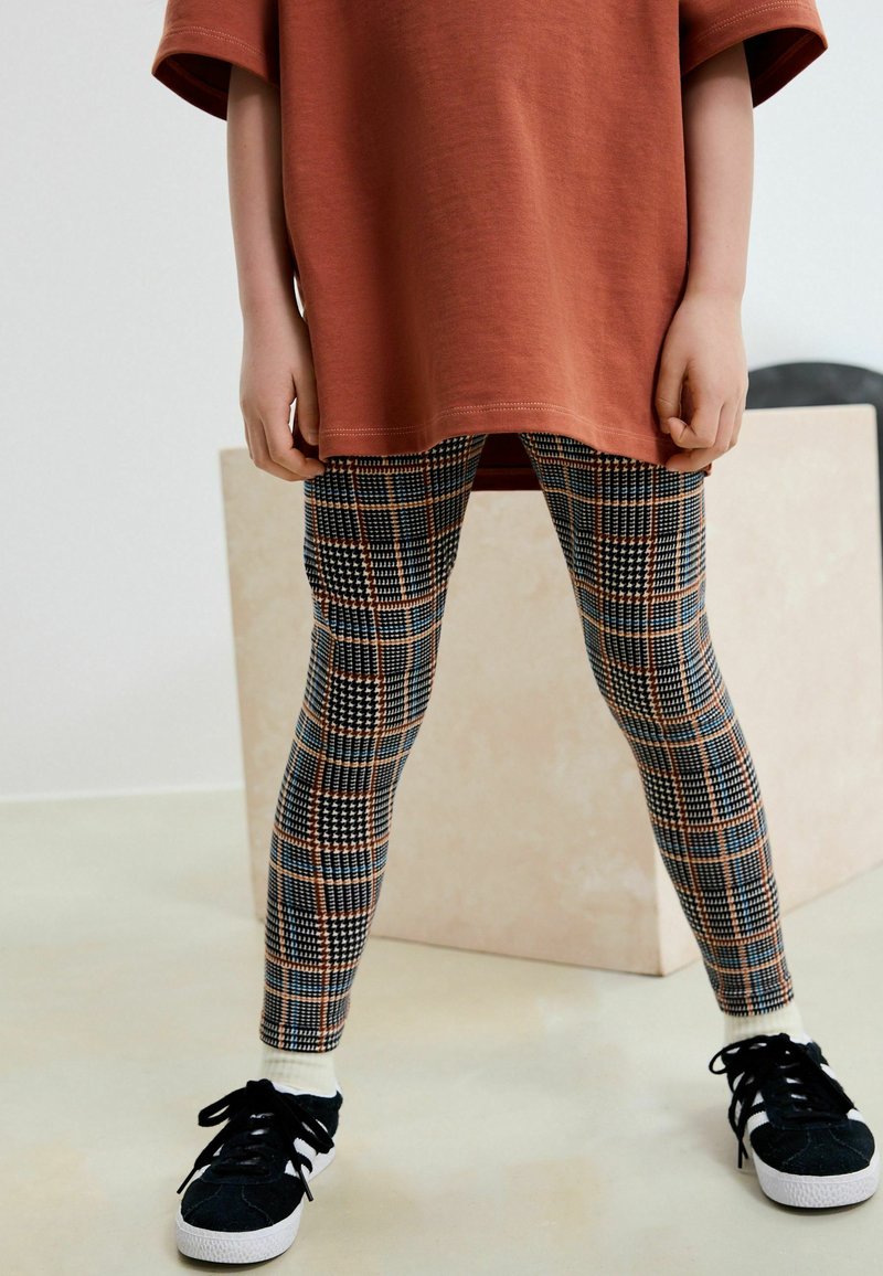 Enfant portant une longue chemise couleur rouille, des leggings à carreaux à motifs, des chaussettes blanches et des baskets noires, debout à l'intérieur.