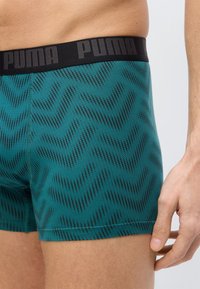 Homme portant un boxer vert sarcelle avec un motif de petits points noirs ondulés et une ceinture noire portant le logo "PUMA" sur le côté.