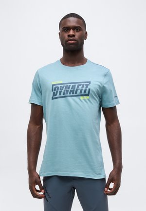 Mladý muž v světlemodrém tričku Dynafit a tmavě šedých šortkách stojí proti jednobílému pozadí.