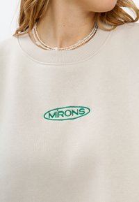 Crème sweatshirt met geribde ronde hals en geborduurd groen "MÍRONS" logo aan de voorkant. Zacht materiaal met een gladde textuur.