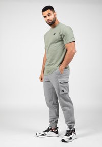 Olijfgroene katoenen t-shirt met een klein logo, gecombineerd met grijze cargo broek met zijzakken en een geborduurd accent. Zwart-witte sneakers.