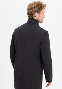 Lange zwarte jas met een hoge kraag, soepele textuur en een op maat gemaakte pasvorm. Heeft zijnaad en een subtiele brandingdetail op de kraag.