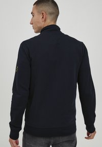 Felpa a maniche lunghe di colore blu navy realizzata in misto cotone. Presenta un colletto alto, polsini e orlo a costine, con un logo ricamato sulla manica.