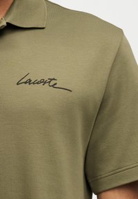 Къса риза с яка в маслиненозелен цвят с черен курсивен надпис "Lacoste" на лявата гърда.