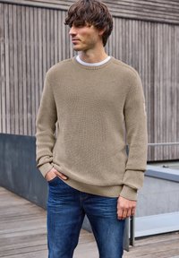 Beige gebreide trui met een gestructureerd patroon, ronde hals en geribbelde manchetten, gecombineerd met blauwe jeans. Buitensetting met een houten achtergrond.
