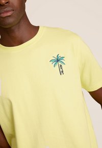 Geel katoenen T-shirt met een klein, multicultureel geborduurd palmbomenontwerp met de tekst "aloha" onder de boom.
