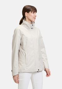 Veste imperméable beige clair avec un col haut, des boutons-pression et des poches latérales ; présente une texture lisse et une silhouette légèrement cintrée.