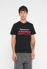 Black cotton T-shirt featuring white and red graphic text "Highperformance 1981 REPLAY" across the chest. Short sleeves and crew neckline.