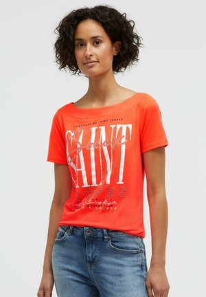 Young woman with curly hair wearing a bright orange "Saint Tropez" printed t-shirt and blue jeans, standing against a plain background.