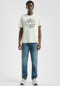 Młody mężczyzna ubrany w biały t-shirt z napisem "Quiet Sense", niebieskie dżinsy i białe trampki, stojący na tle jasnego, jednolitego tła.