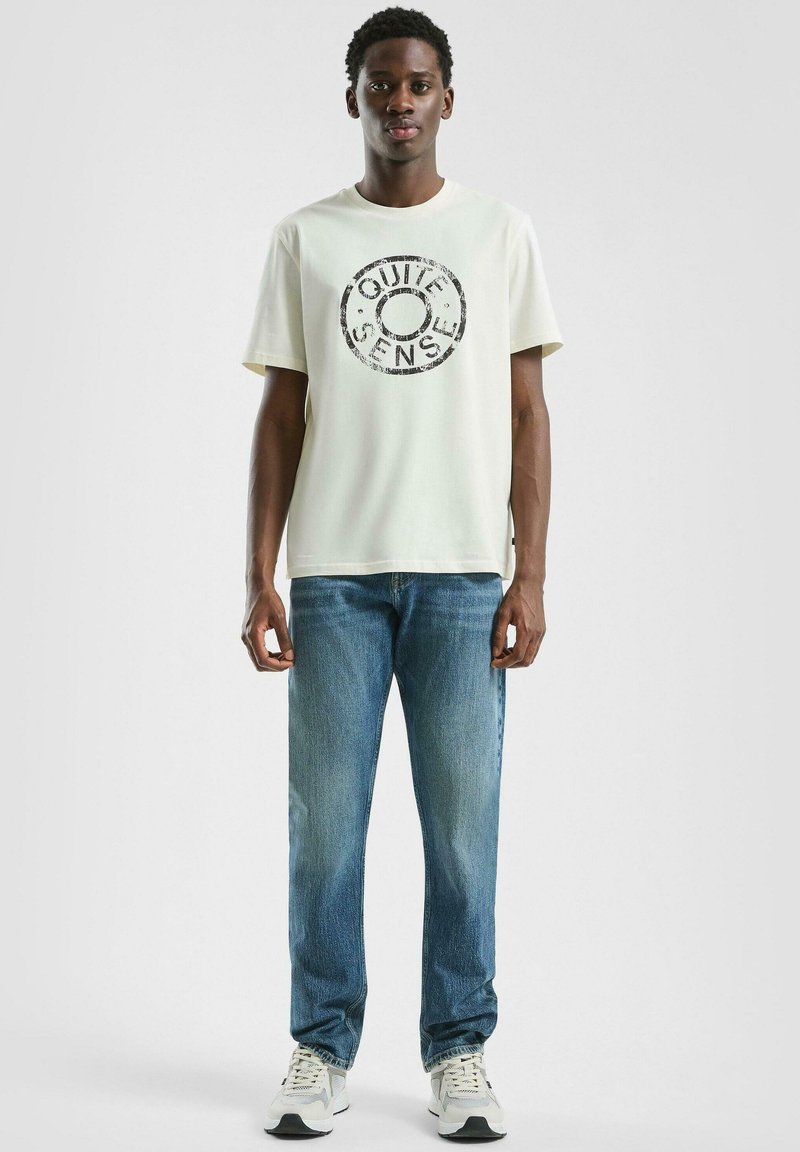 Młody mężczyzna ubrany w biały t-shirt z napisem "Quiet Sense", niebieskie dżinsy i białe trampki, stojący na tle jasnego, jednolitego tła.