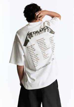 Person med kort mørkt hår iført en hvid Metallica tour t-shirt med opremsning af europæiske byer og datoer fra 1986, står med hånden på nakken.