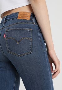 Tmavě modré džíny z denimu s těsným střihem, na zadním pase ozdobené hnědou koženou etikettou a charakteristickým zakřiveným prošíváním zadních kapes.