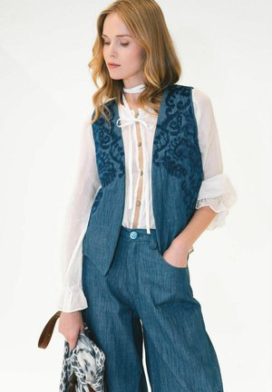 Giovane donna che indossa una camicetta bianca con fiocco, un gilet blu con fantasia, pantaloni blu, che tiene una sciarpa con fantasia e una tracolla marrone.