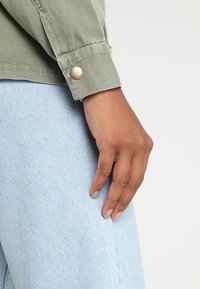 Green canvas shirt cuff with gold snap button, paired with light blue denim. The fabric has a smooth texture with visible stitching.