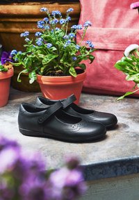 Scarpe Mary Jane in pelle nera con punta arrotondata, cinturino sulla pianta del piede e dettagli scallopati; collocate su uno sfondo di fiori colorati in vaso.