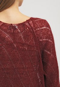 Burgunderfärgad stickad tröja med diamantsmönster och öppen vävstruktur. Har rundad halsringning och knappdetalj på baksidan.