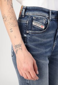 Tamno plave traperice s izbljedjelošću, dizajn s pet džepova i oznakom marke na stražnjoj strani. Tetovaža je vidljiva na zapešću.