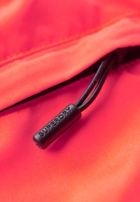 FelOranje stof met een gladde textuur heeft een zwarte trekkoord en een label met de tekst "SUPERDRY" nabij een ritssluiting.