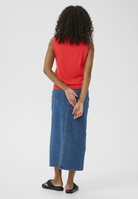 Un haut sans manches rouge, une jupe maxi en denim, des sandales noires. La jupe présente une fente à l'arrière et des coutures visibles sur les poches. Cheveux bouclés, vus de dos.